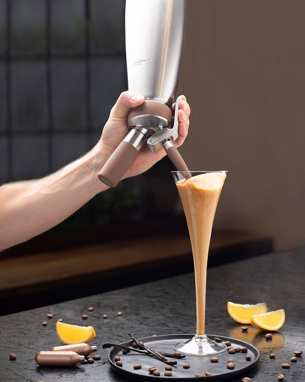 Jemand sprüht mit einem iSi Nitro Espresso Martini in ein Glas, daneben iSi Charge Kapseln und Orangen. 