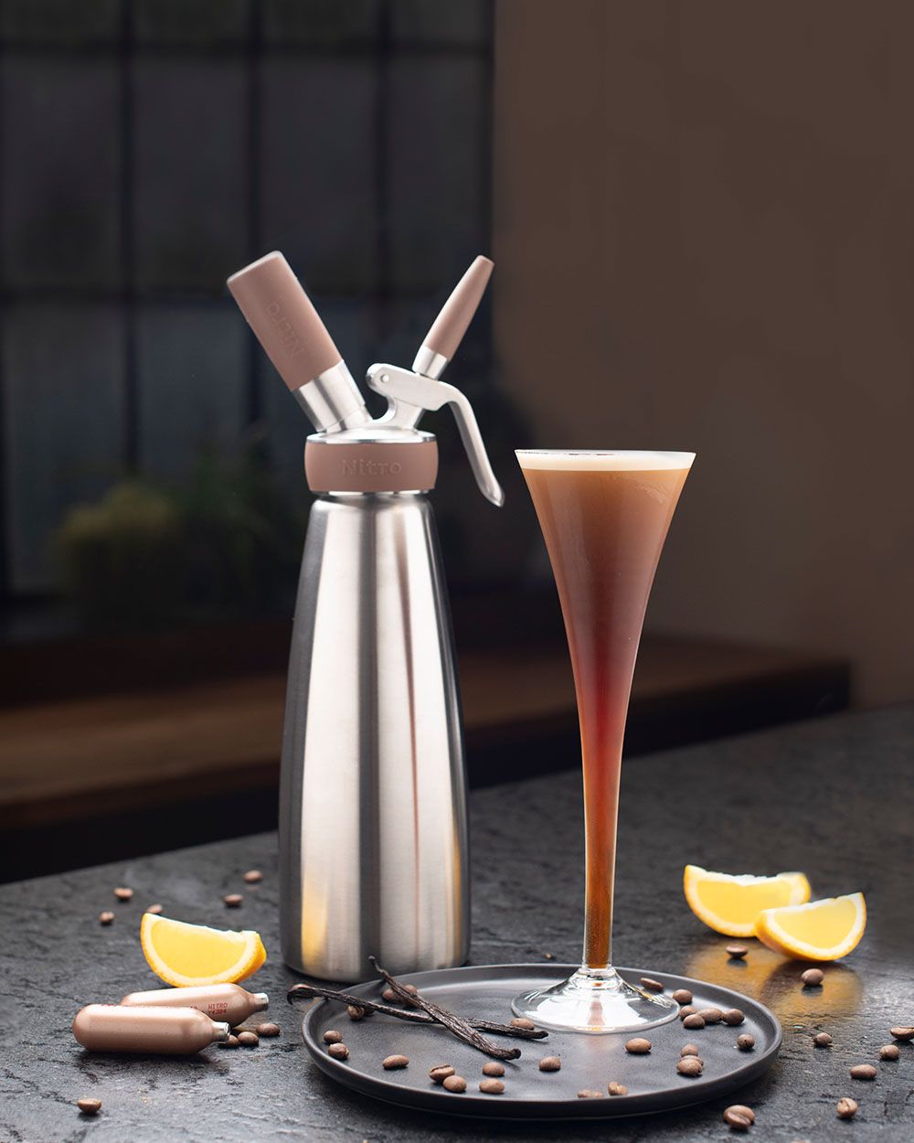 Ein Glas mit Nitro Espresso Martini, daneben ein iSi Nitro, Orangenscheiben, iSi Charge Kapseln und Kaffeebohnen. 