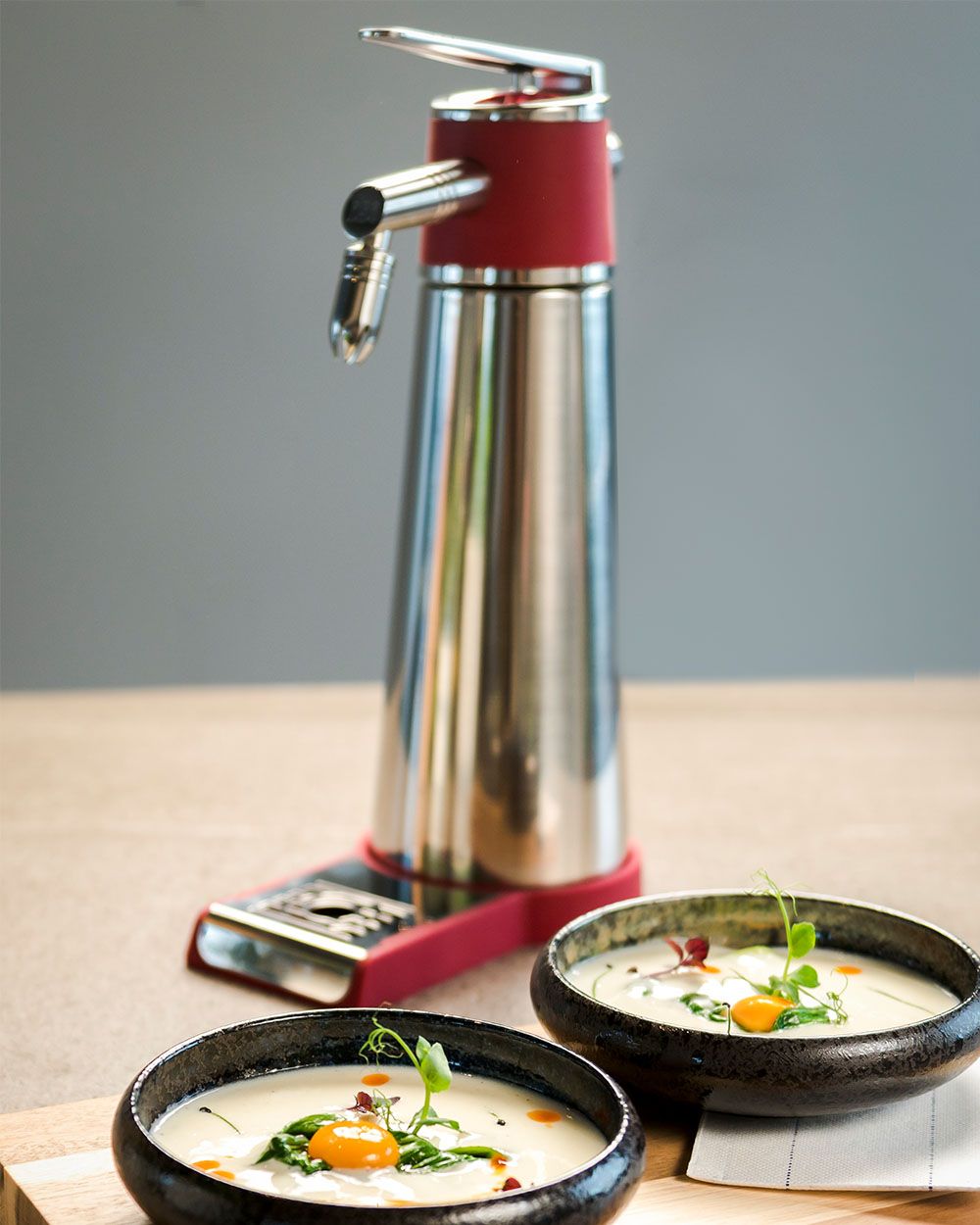 Zwei Schalen mit cremiger Suppe, garniert mit frischen Kräutern und Tomaten, stehen vor einem iSi Whip mit rotem Sockel.