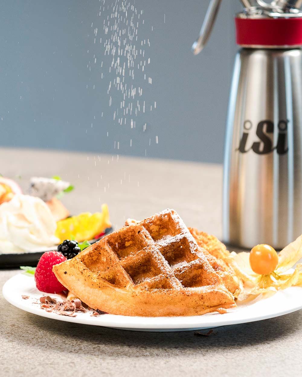 Auf dem Bild wird Puderzucker über eine frische Waffel mit Beeren und Physalis gestreut, im Hintergrund steht ein iSi Gourmet Whip.