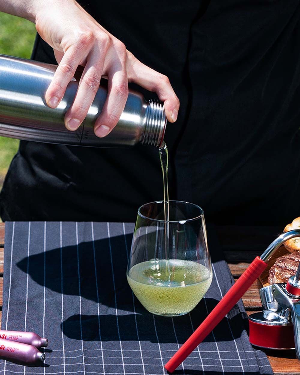 Jemand gießt mediterranes Öl aus einem iSi Gourmet Whip in ein Glas, daneben iSi Charge Kapseln. 