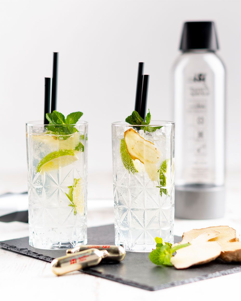 Zwei Longdrinkgläser mit sprudelndem Getränk, Limettenscheiben, Minze und Apfelchips stehen auf Schieferplatten, daneben liegen goldene iSi Kapseln und im Hintergrund ein iSi Sodamaker.