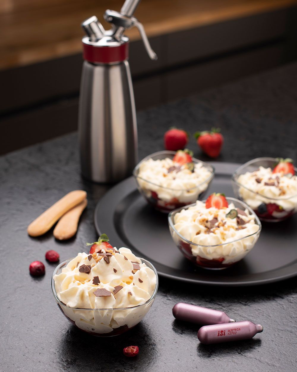 Mehrere Schüsseln mit Tiramisu und Beeren, daneben iSi Charge Kapseln und um Hintergrund mehr Beeren, Biskotten und ein iSi Gourmet Whip.