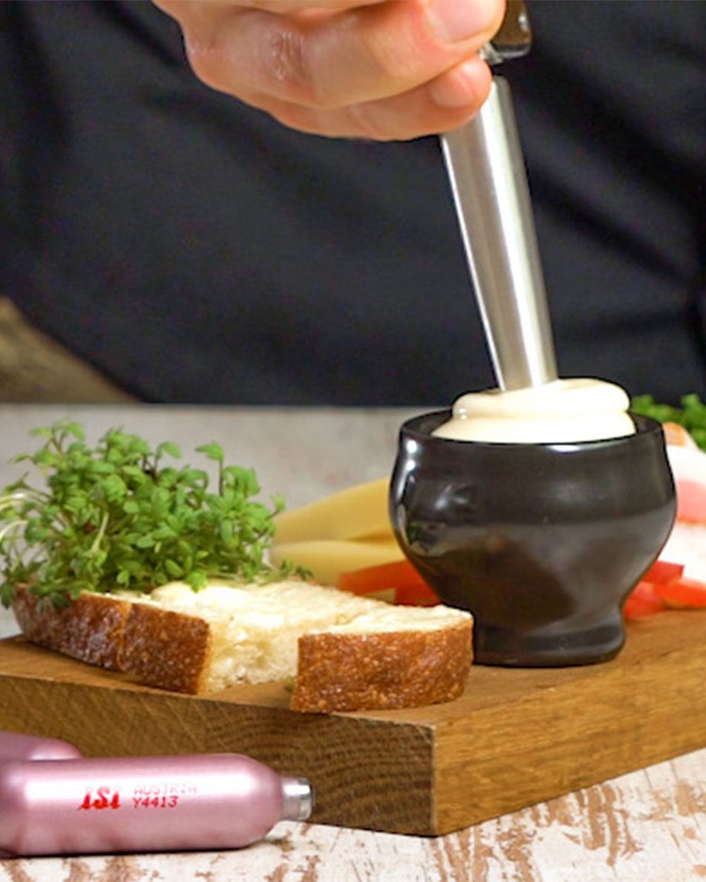 Jemand nutzt ein iSi Gourmet Whip, um Eiaufstrich in eine kleine Schüssel zu sprühen. Daneben liegt ein Stück Brot mit Kräutern und zwei iSi Charge Kapseln.