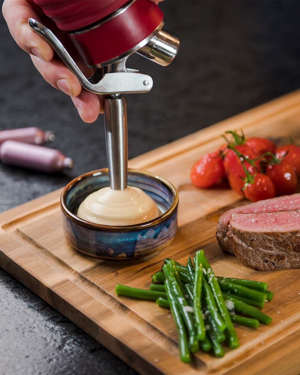 Jemand nutzt einen Gourmet Whip um Cheddar Hollandaise Sauce in eine Schüssel zu sprühen. Auf dem selben Brett liegen Gemüse und Fleisch. 