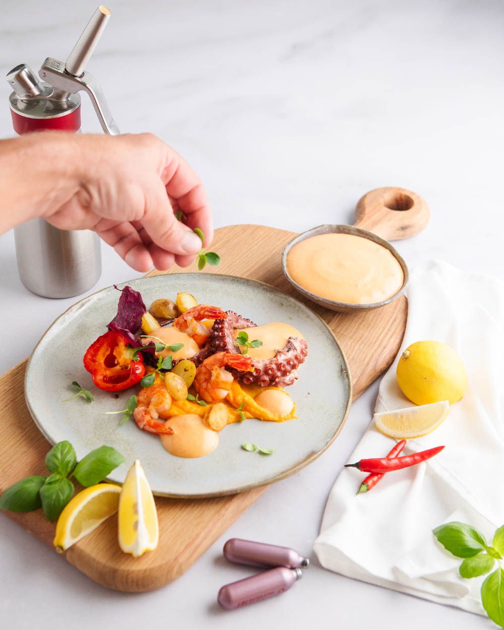 Jemand gibt Gewürze auf einen Teller mit Meeresfrüchten und Chilli Mayonnaise. Daneben Zitronen, mehr Sauce und zwei iSi Charge Kapseln. 