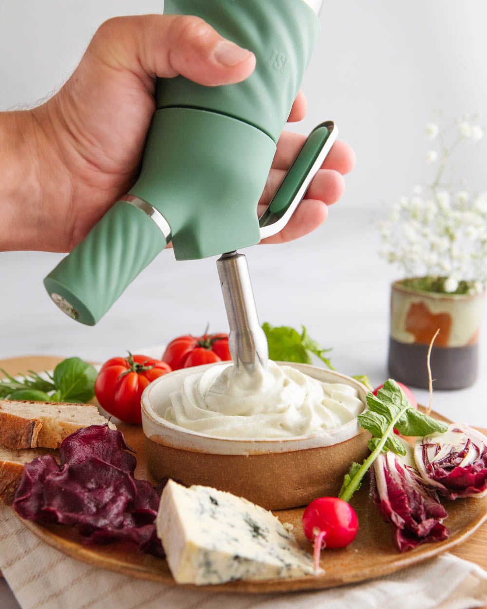 Eine Hand spritzt mit einem iSi Whip cremigen Dip in eine Schale, die zusammen mit Käse, Radieschen, Tomaten, Brot und Gemüsechips auf einer Servierplatte angerichtet ist.