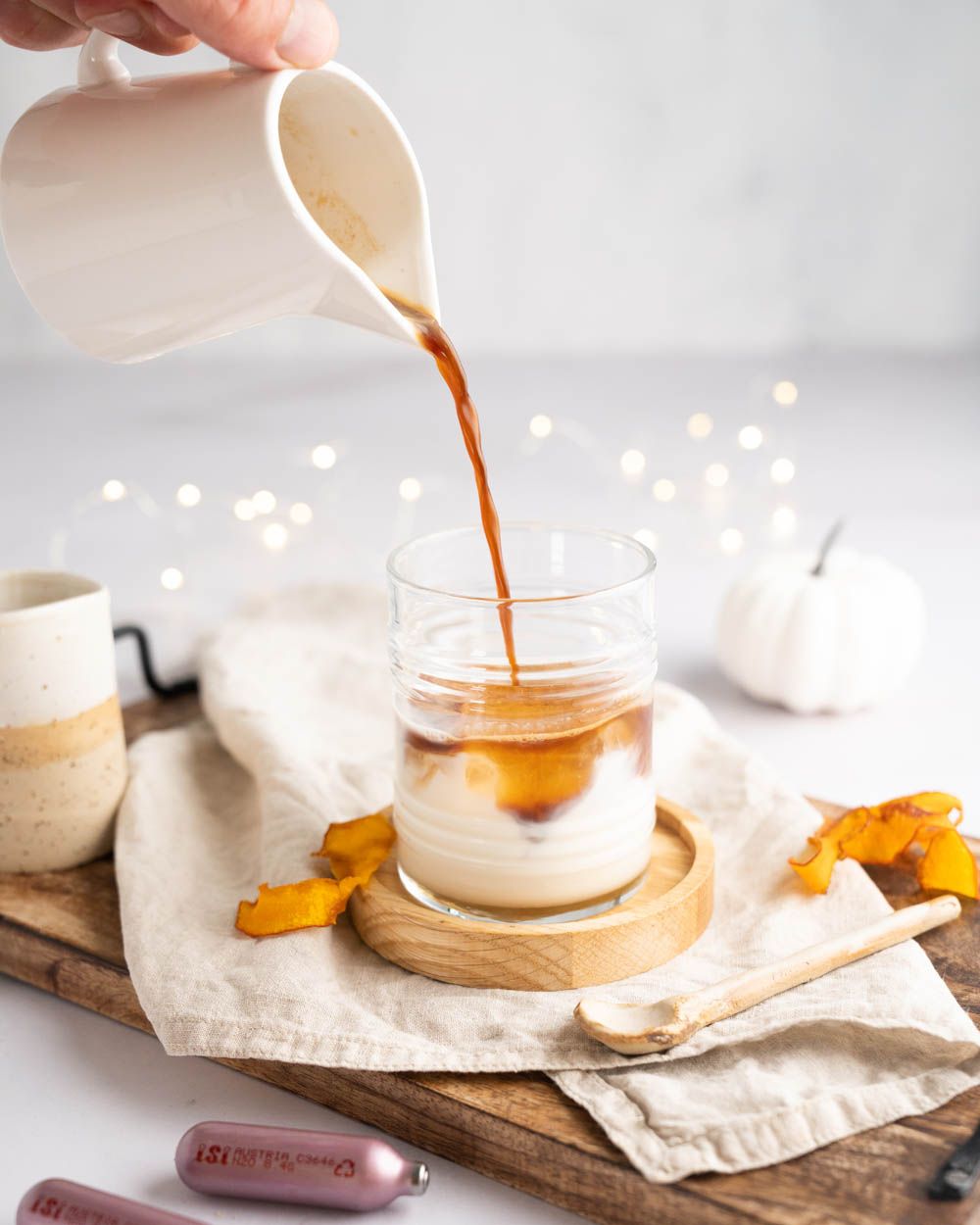 Jemand gießt Kaffee in ein Glas mit Milch, daneben Kürbis und iSi Charge Kapseln. 