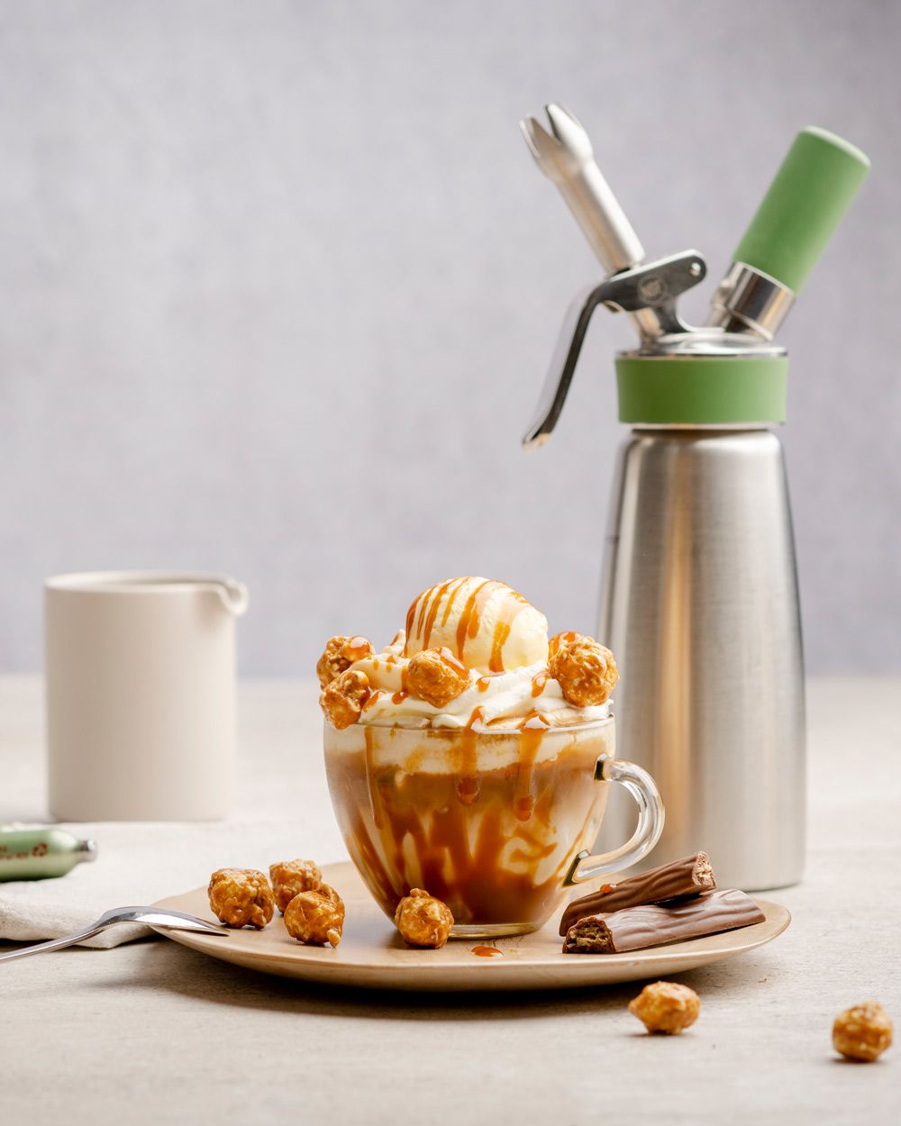 Auf einem Teller steht ein fertiger Affogato Kaffee, daneben liegen Haselnüsse und Schokoriegel und im Hintergrund steht ein iSi Green Whip. 