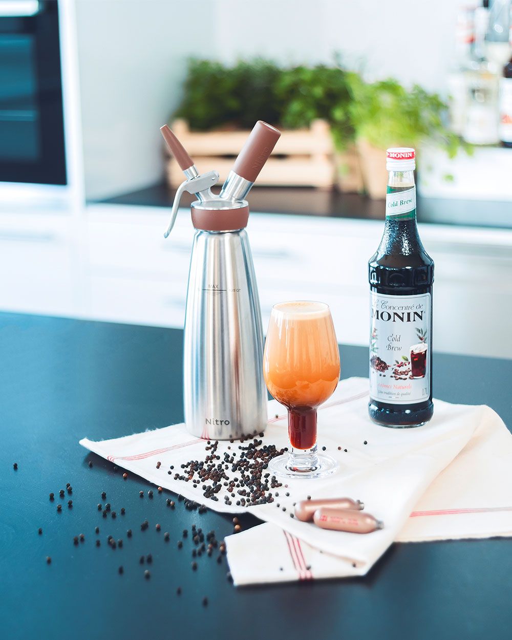 Ein Glas mit Dark Brew, daneben ein iSi Nitro, eine Flasche Monin Sirup und iSi Charge Kapseln. 