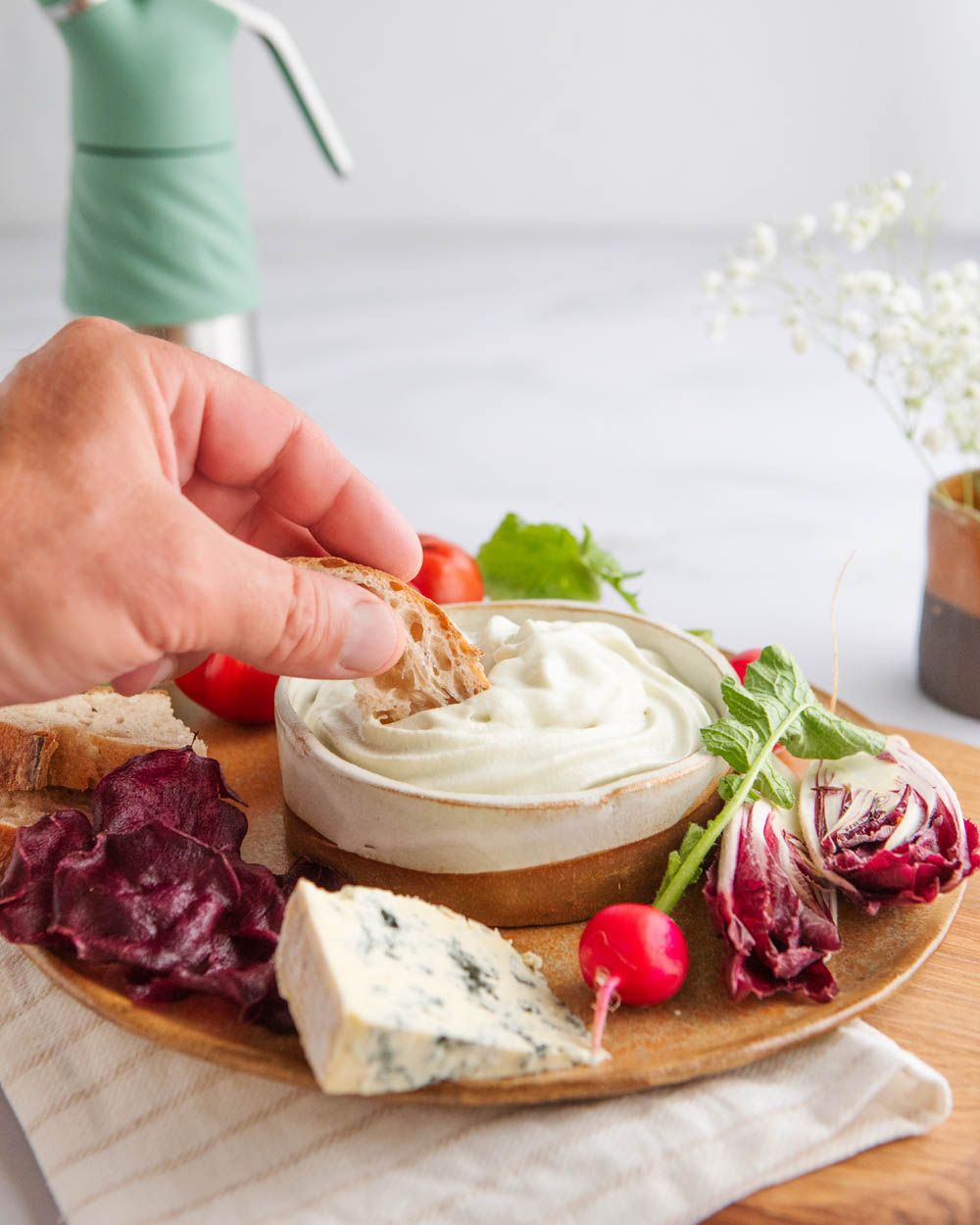Eine Hand tunkt ein Stück Brot in eine Schale mit cremigem Dip, die auf einer Servierplatte mit Käse, Radieschen, Tomaten und Gemüsechips angerichtet ist, im Hintergrund steht ein iSi Whip.