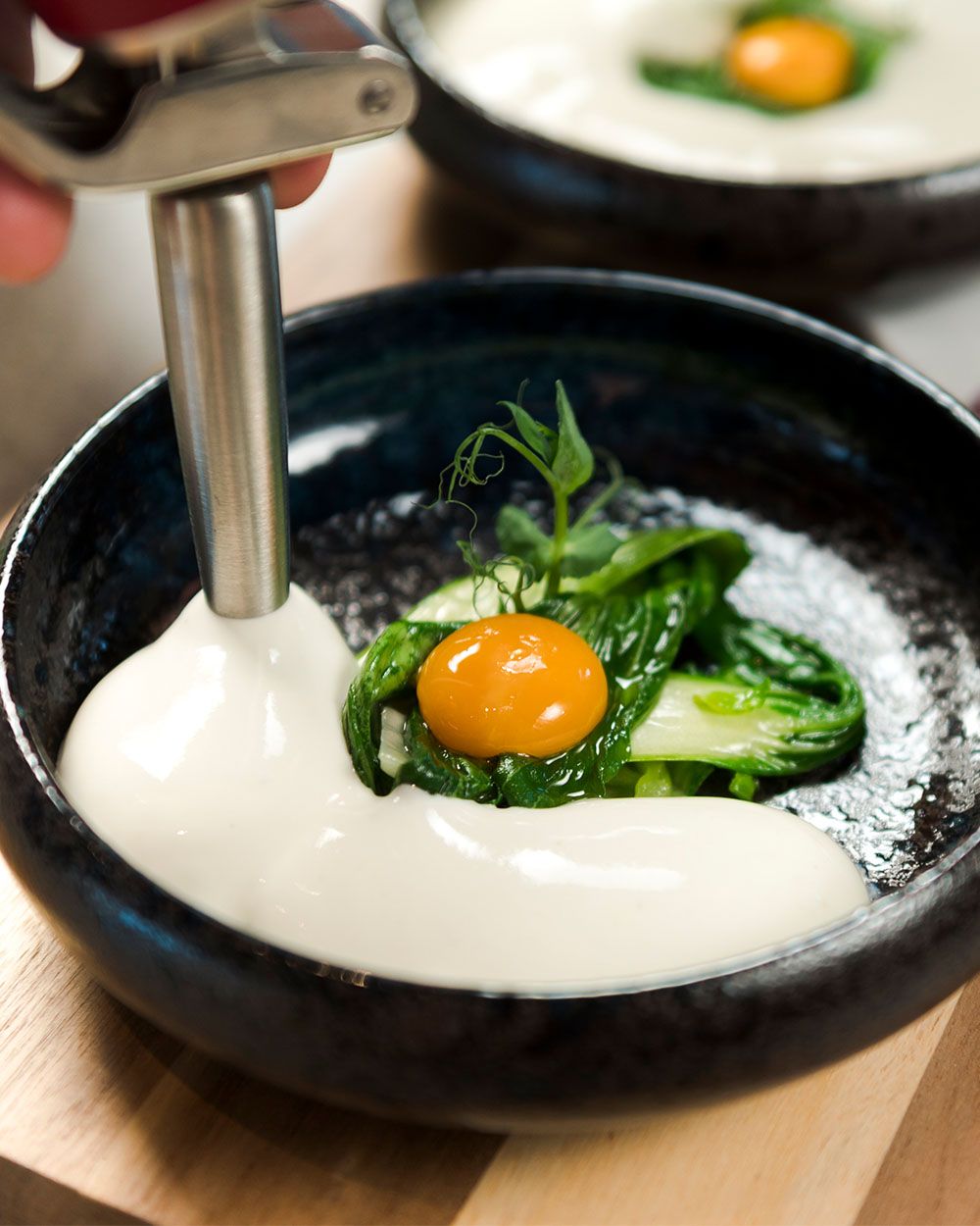 Eine cremige Sauce wird mit einem iSi Whip in eine Schale gefüllt, in der Pak Choi und ein Eigelb als Garnitur angerichtet sind.