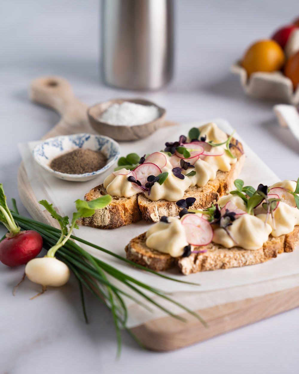 Zwei Brote mit Eiaufstrich und Kräutern, daneben Schüsseln mit Gewürzen und Radieschen. Im Hintergrund ein iSi Gourmet Whip, iSi Charge Kapseln und bunte Eier. 