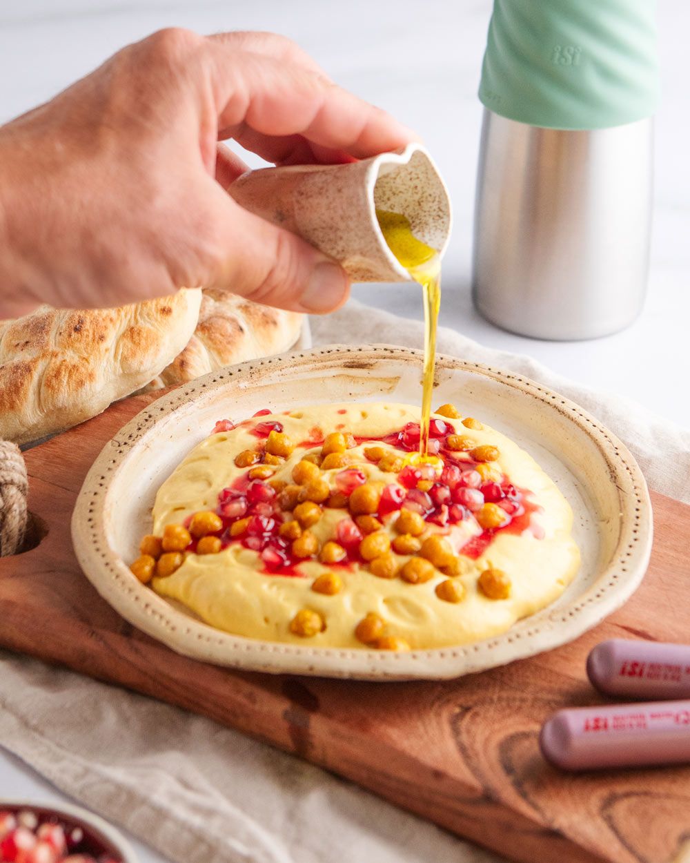 Jemand gießt Öl auf einen Teller mit Kürbishummus, daneben Pitabrot und zwei iSi Charge Kapseln. Im Hintergrund ein iSi Creative Whip. 