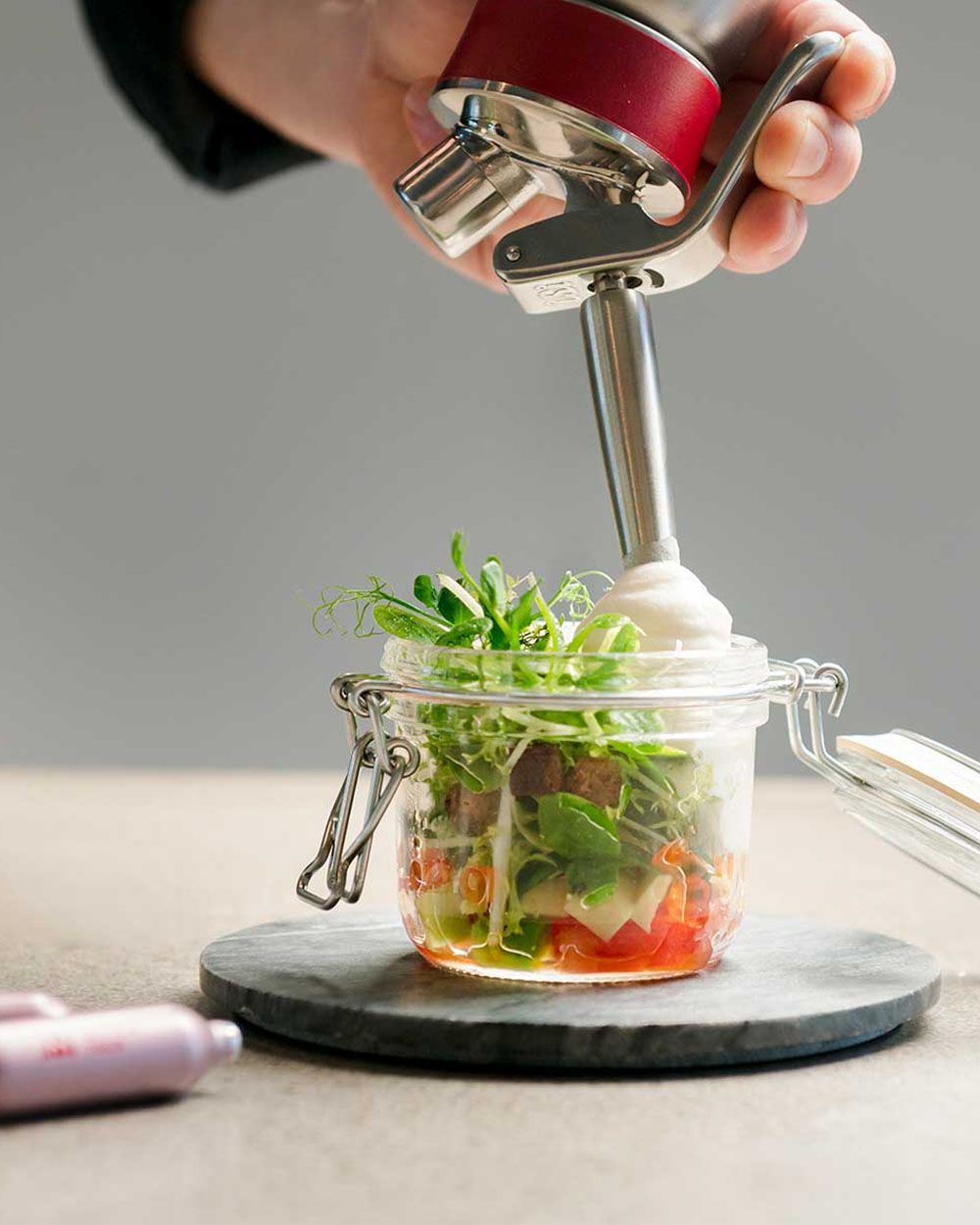Jemand sprüht mit einem iSi Gourmet Whip Forellen-Mousse in ein Glas mit Gemüse, daneben iSi Charge Kapseln. 