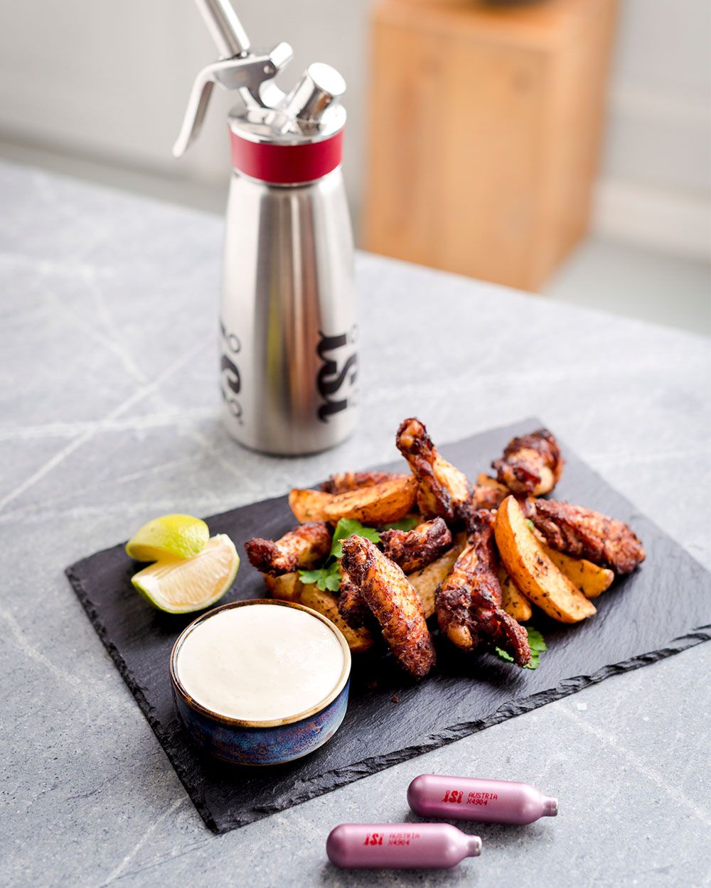 Auf dem Bild steht ein iSi Gourmet Whip mit rotem Ring hinter einem Servierbrett mit Chicken Wings, Kartoffelspalten, Limettenstücken und einem Dip.