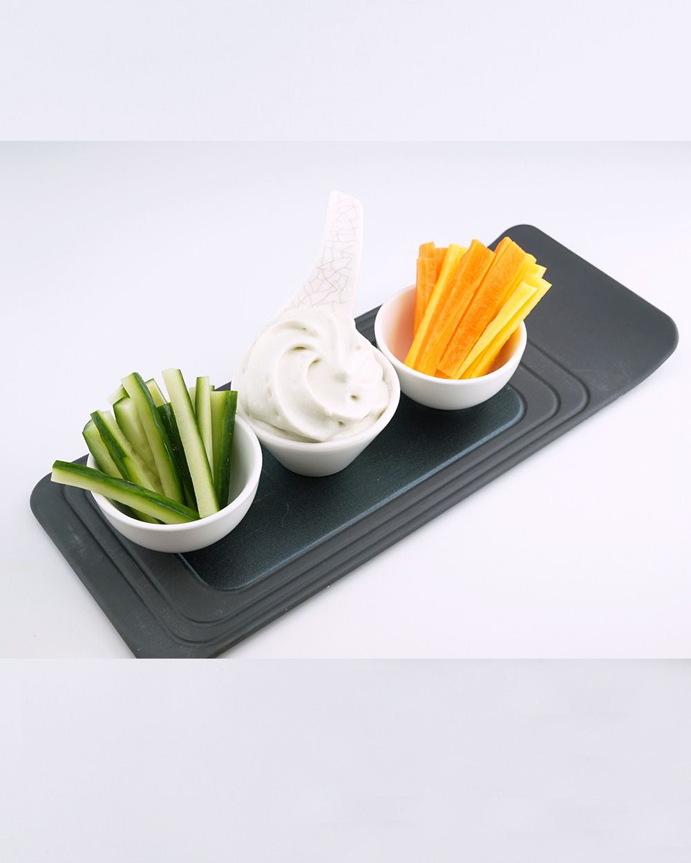 Drei kleine Schälchen mit Gurkensticks, cremigem Dip und Karottensticks stehen dekorativ auf einer schwarzen Servierplatte.