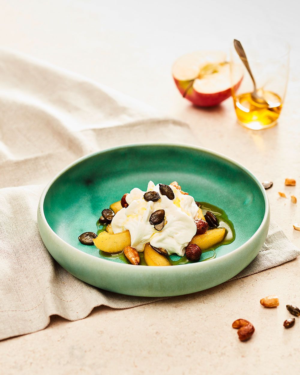 Ein Dessertteller mit geschmorten Apfelspalten, Nüssen, Rosinen und cremigem Topping ist auf einem grünen Teller angerichtet, im Hintergrund stehen ein Glas mit Honig und ein halbierter Apfel.