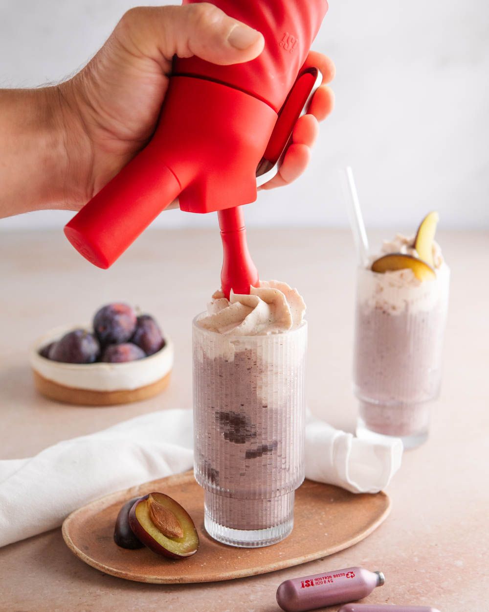 Jemand sprüht mit einem iSi Creative Whip Zimt-Topping auf ein Pflaumen-Milch-Getränk. Davor liegen mehr Pflaumen und eine iSi Charge Kapsel.