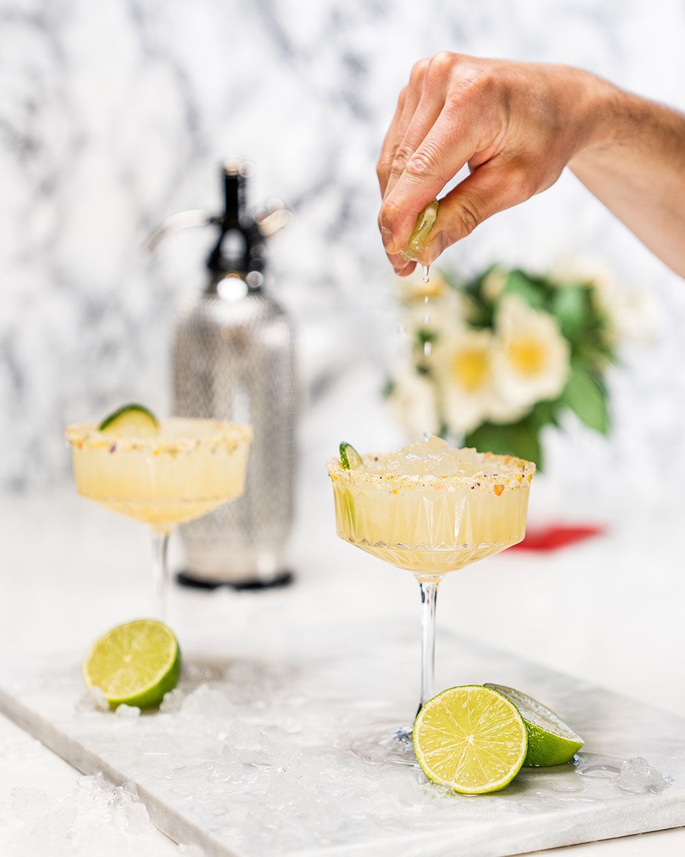 Jemand presst Limettensaft in ein Glas mit Holunder-Minz-Limonade, daneben Limetten und im Hintergrund Blumen und ein iSi Sodamaker. 