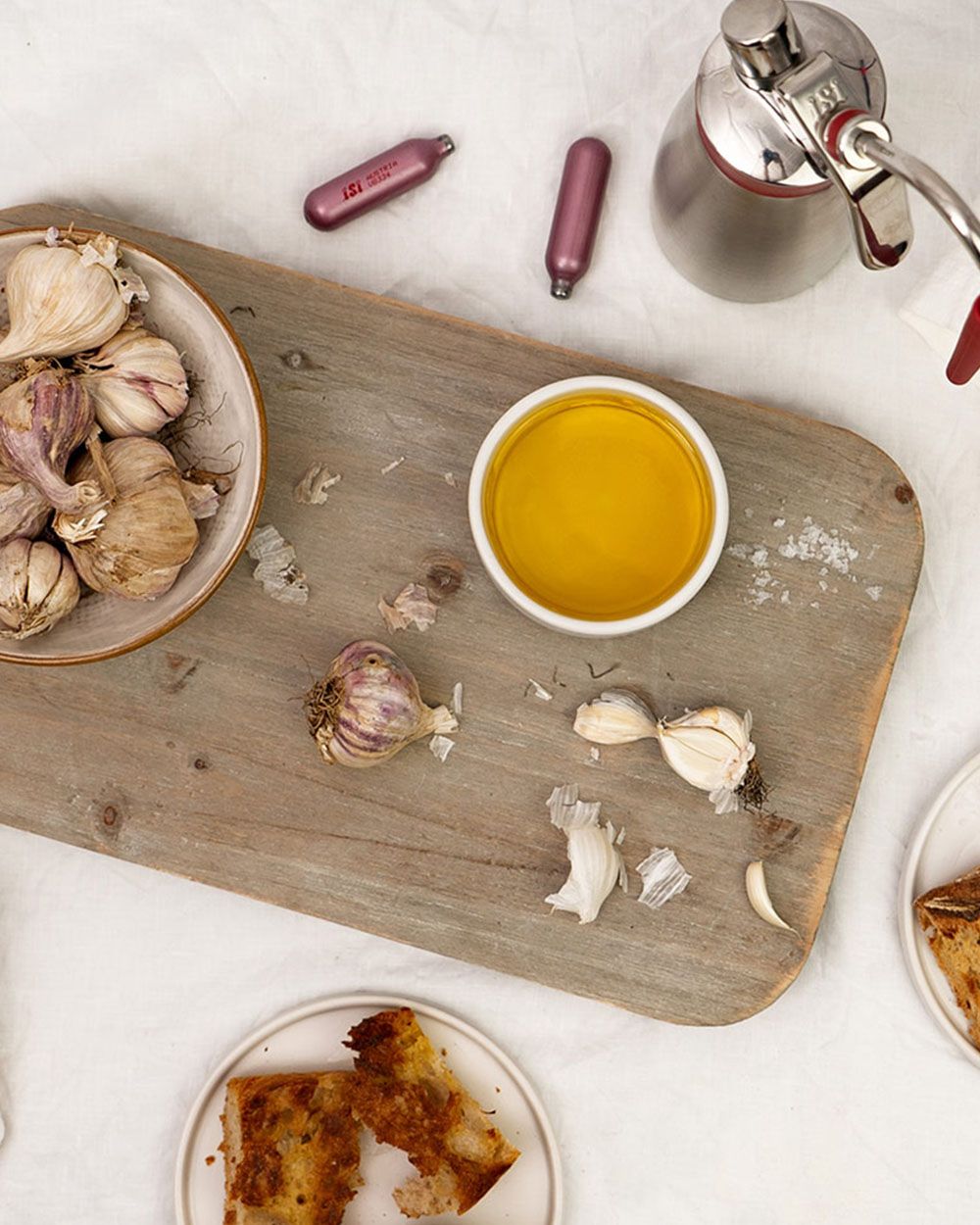 Ein Brett auf dem Knoblauch und eine Schüssel mit Knoblauchsauce sind, daneben iSi Charge Kapseln und ein iSi Gourmet Whip. 