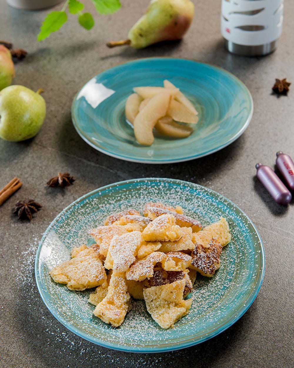 Ein Teller mit Rahmschmarren, dahinter ein Teller mit gekochten Birnen. Daneben Birnen, Zimt und zwei iSi Charge Kapseln. 