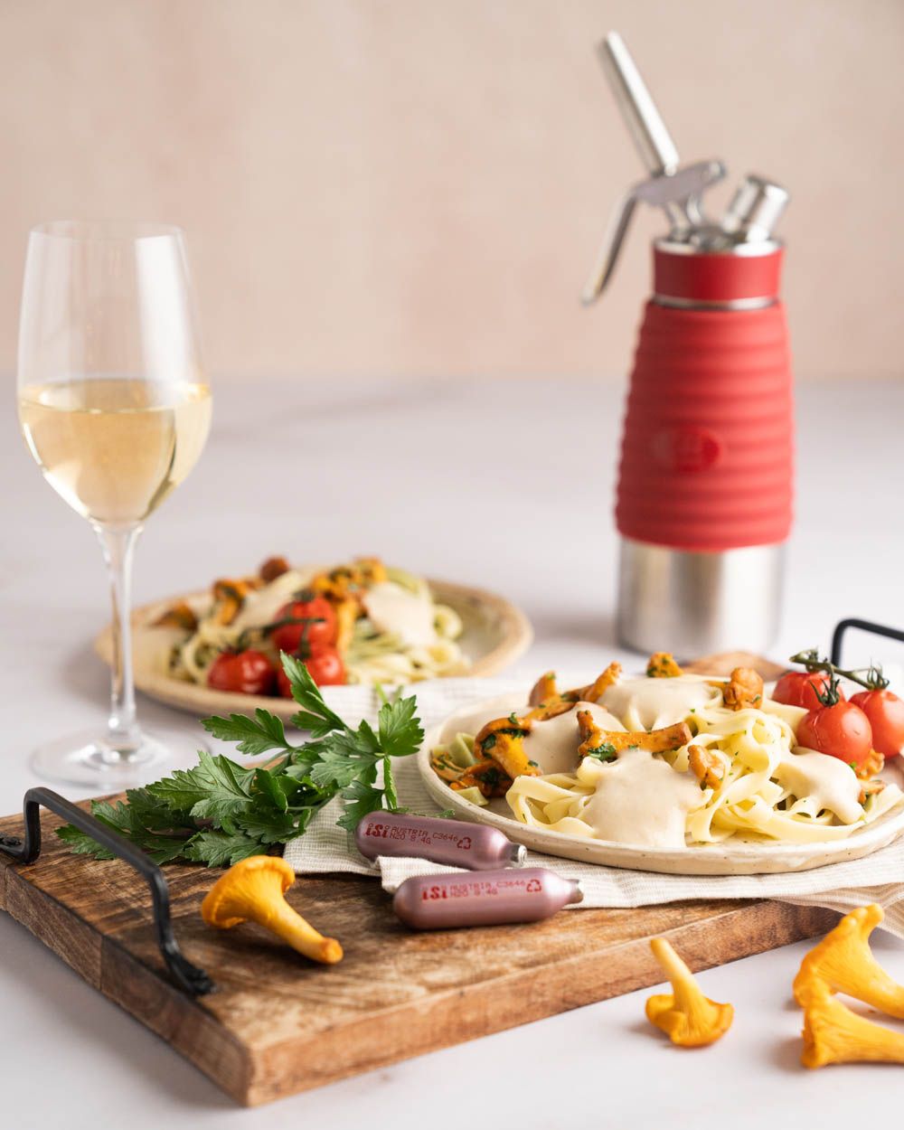 Zwei Teller mit Pasta und Pilzrahmsauce. Daneben Pilze, Kräuter, ein Glas Wein und zwei  iSi Charge Kapseln. Im Hintergrund ein iSi Gourmet Whip. 