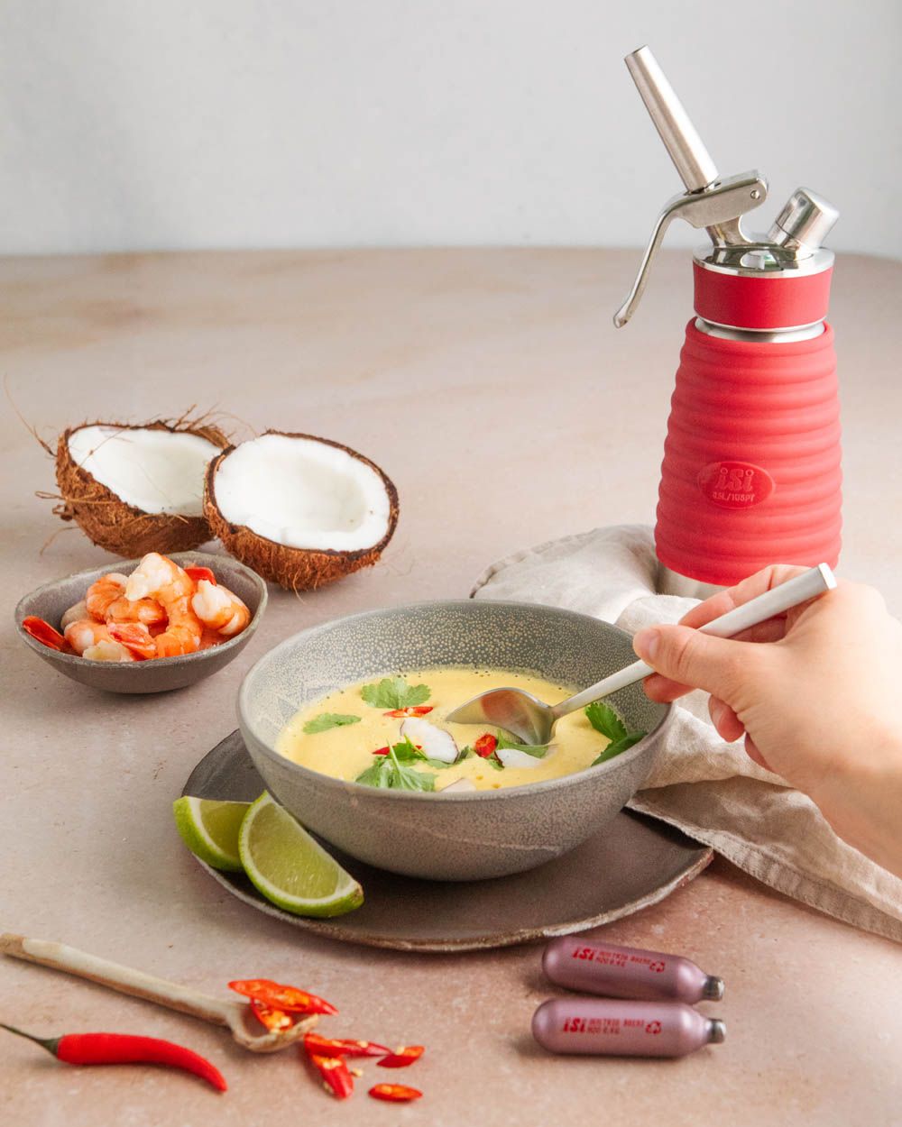 Jemand löffelt thailändische Kokos-Suppe, daneben Kokosnüsse, Gemüse, iSi Charge Kapseln und ein iSi Gourmet Whip. 