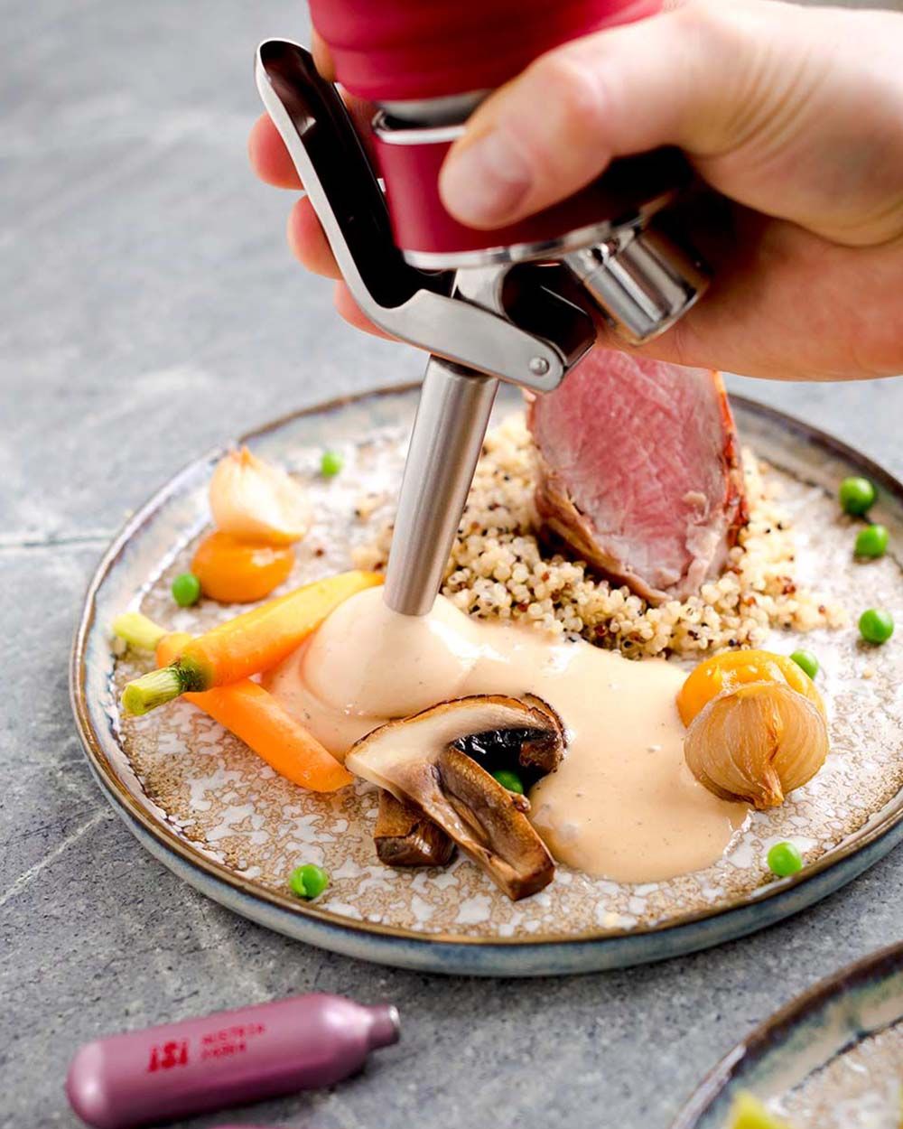 Jemand sprüht mit einem iSi Gourmet Whip Wacholdercremesauce auf einen Teller mit Fleisch und Gemüse. Davor liegt eine iSi Charge Kapsel. 