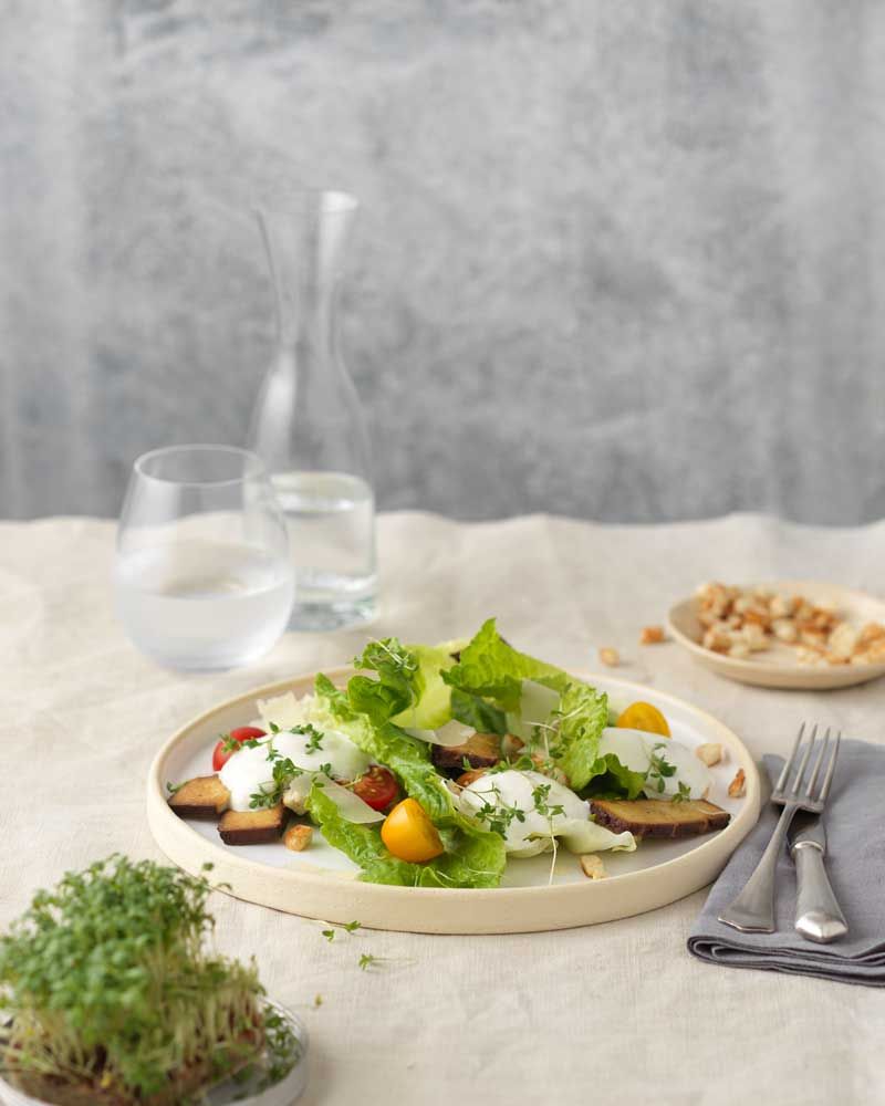 Auf dem Bild ist ein frischer Salat mit Romanasalat, Tomaten, Croûtons, Parmesan und Kräutern auf einem Teller angerichtet.