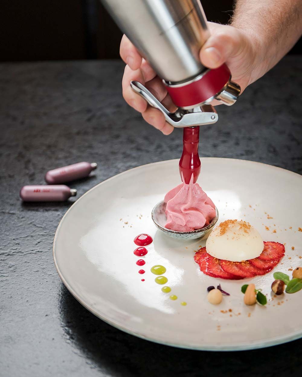 Jemand sprüht mit einem iSi Gourmet Whip Himbeer-Espuma auf einen Teller. Daneben iSi Charge Kapseln. 