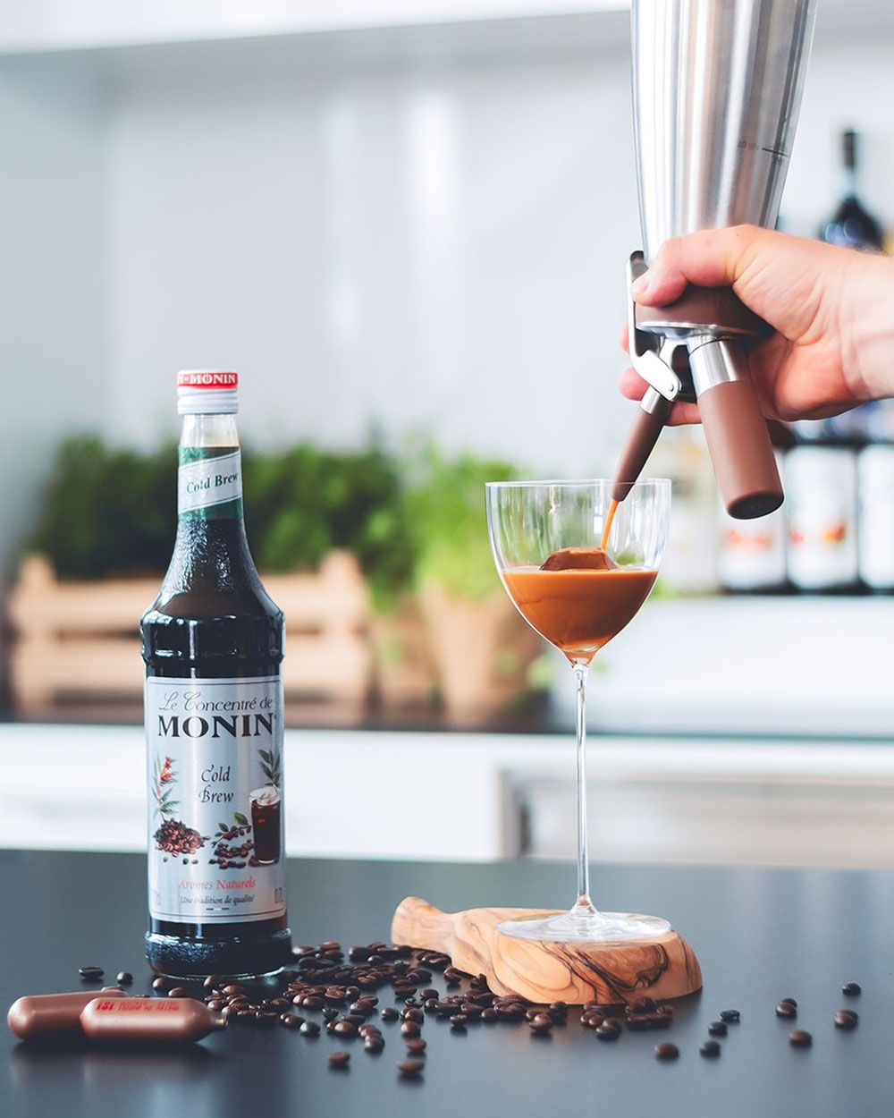 Jemand sprüht aus einem iSi Gourmet Whip Martinicreme in ein Glas, daneben eine Flasche Sirup und iSi Charge Kapseln. 