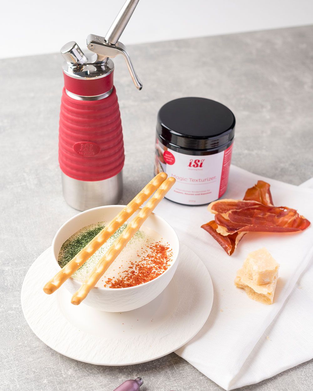 Eine Schale mit cremiger Suppe, dekoriert mit Brotstangen und Gewürzen, steht neben einem iSi Whip, einem iSi Magic Texturizer sowie Parmesanstücken und Schinkenchips.