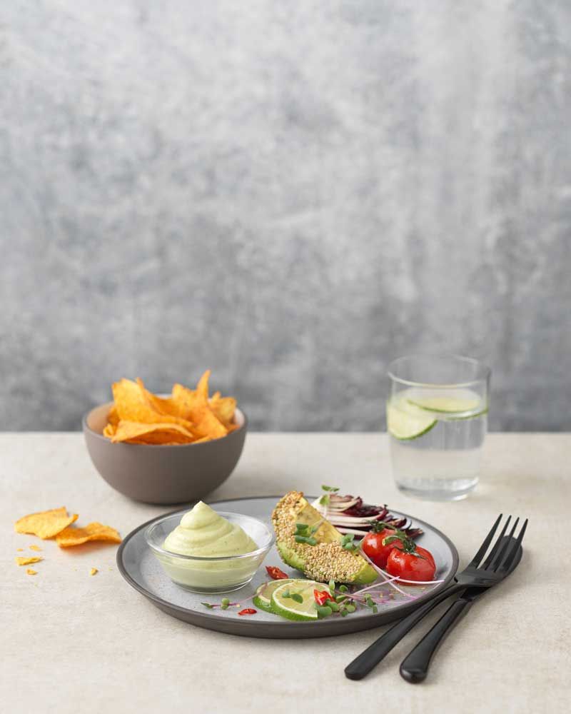 Ein Teller mit Guacomole Dip, daneben Gemüse und Besteck. Im Hintergrund eine Schüssel mit Nachos und ein Glas Wasser. 