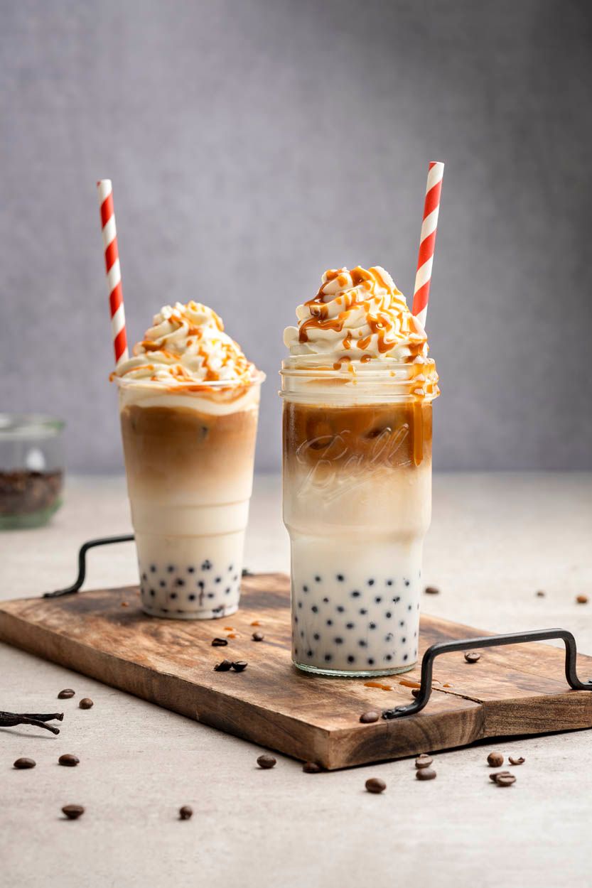 Zwei Iced Bubble Coffees mit Sahneschaum auf einem Holztablett mit Griffen. Daneben liegen Kaffeebohnen. 