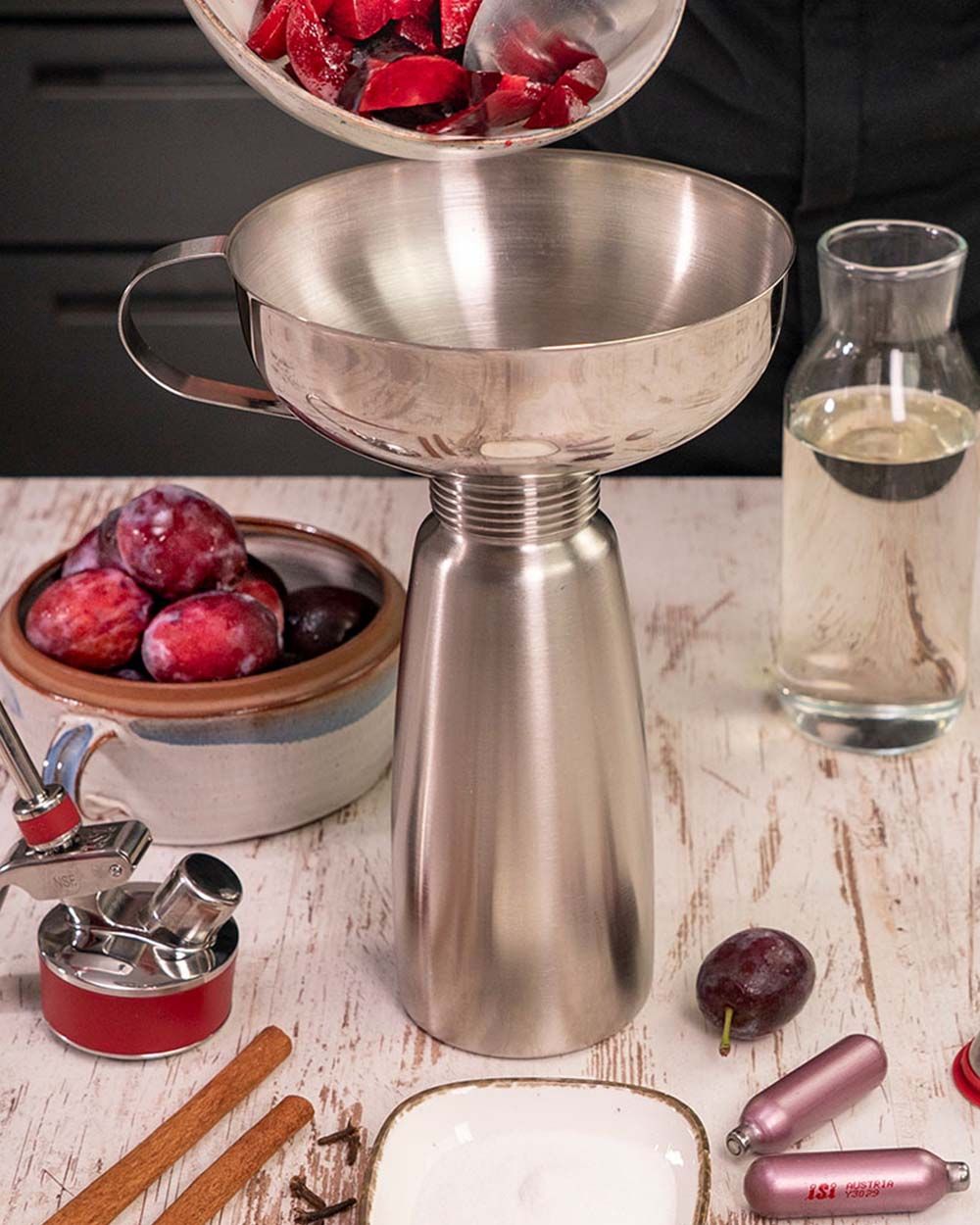 Jemand gibt Pflaumen in ein iSi Gourmet Whip um Pflaumensirup herzustellen, daneben weitere Pflaumen, Wasser und iSi Charge Kapseln. 