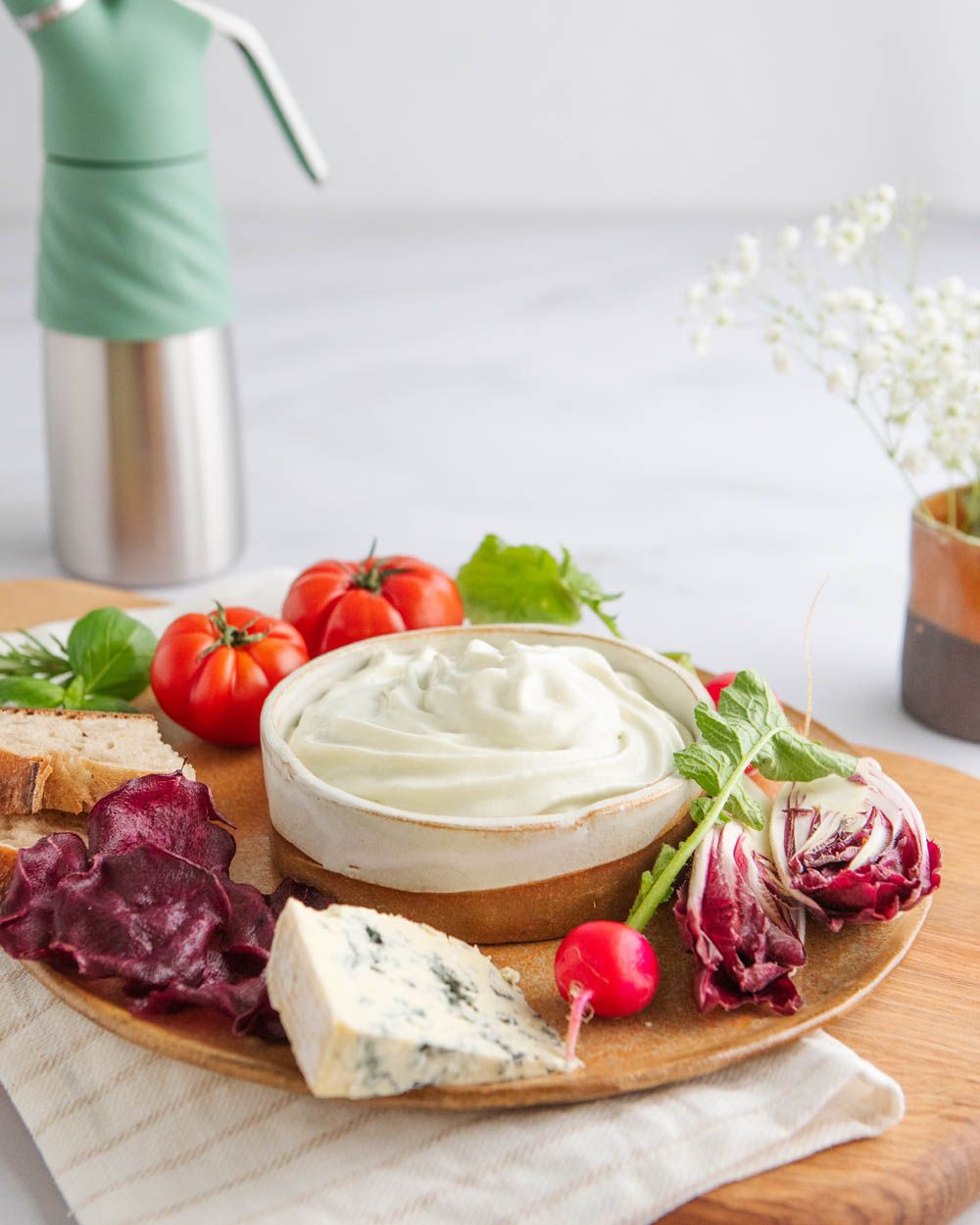 Eine Servierplatte mit cremigem Dip, Blauschimmelkäse, Radieschen, Tomaten, Brot und Gemüsechips steht auf einem Tuch, im Hintergrund ist ein iSi Whip mit grünem Silikonschutz zu sehen.