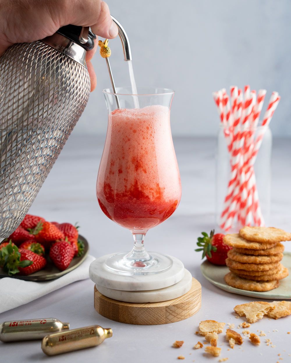Ein sprudelnder Erdbeer-Drink wird mit einem iSi Sodamaker in ein Glas gefüllt, daneben stehen frische Erdbeeren, Kekse, goldene iSi Kapseln und ein Glas mit rot-weißen Strohhalmen.