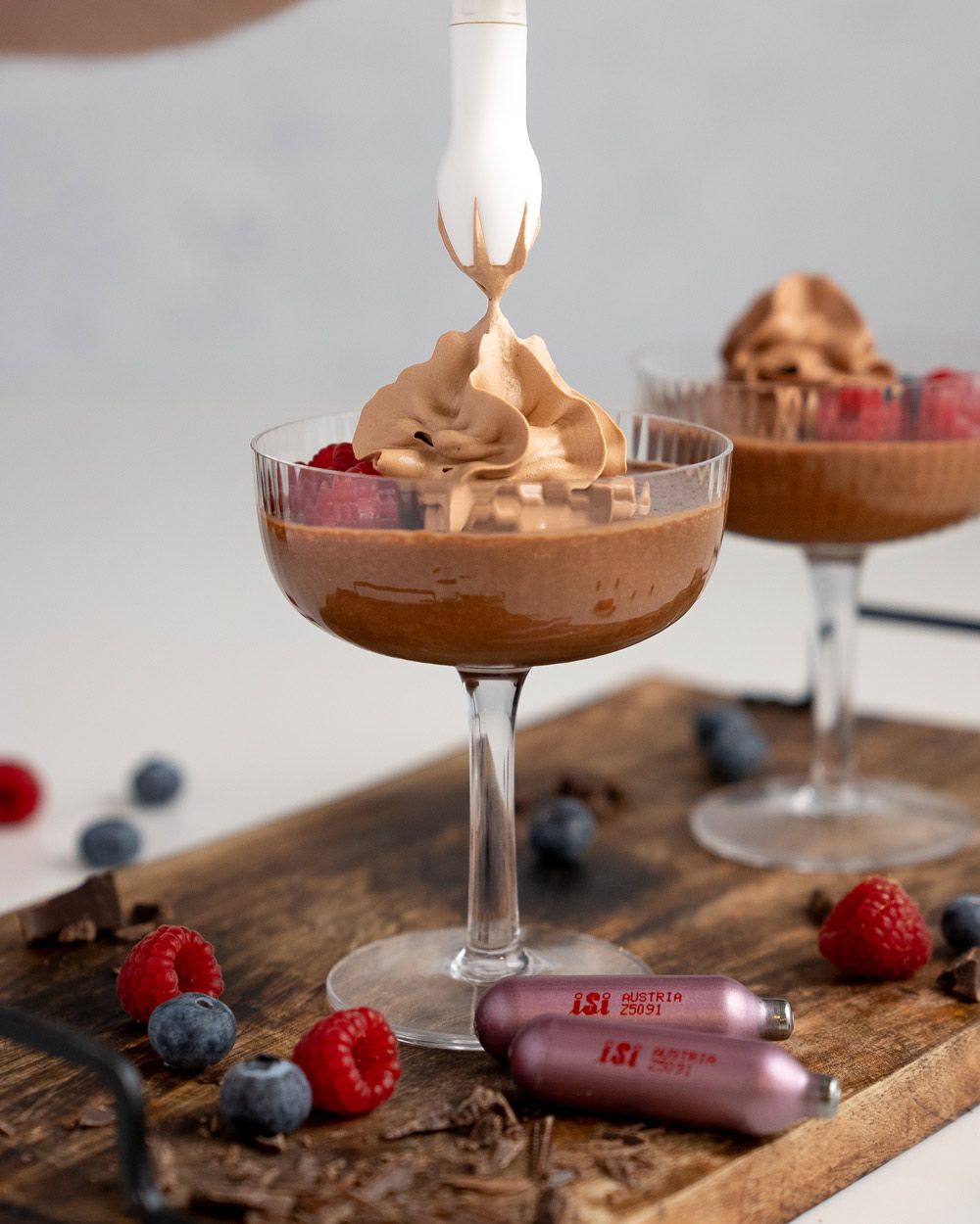 Jemand sprüht Schokomousse in ein Glas, daneben iSi Charge Kapseln, frische Beeren und ein weiteres Glas mit Schokomousse.