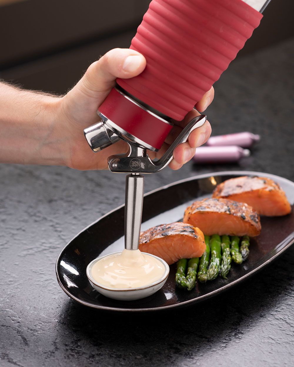 Jemand sprüht mit einem iSi Gourmet Whip Hollandaise Sauce in eine Schüssel, daneben auf dem Teller sind Spargel und Lachs. 