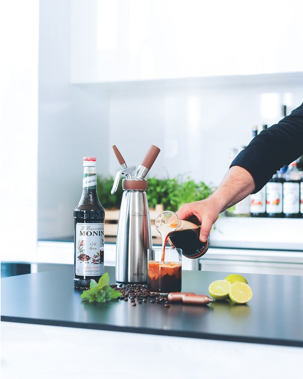 Jemand gießt Hibiskus Kombucha in ein Glas, daneben Limetten, iSi Charge Kapseln, Monin Sirup und ein iSi Nitro. 