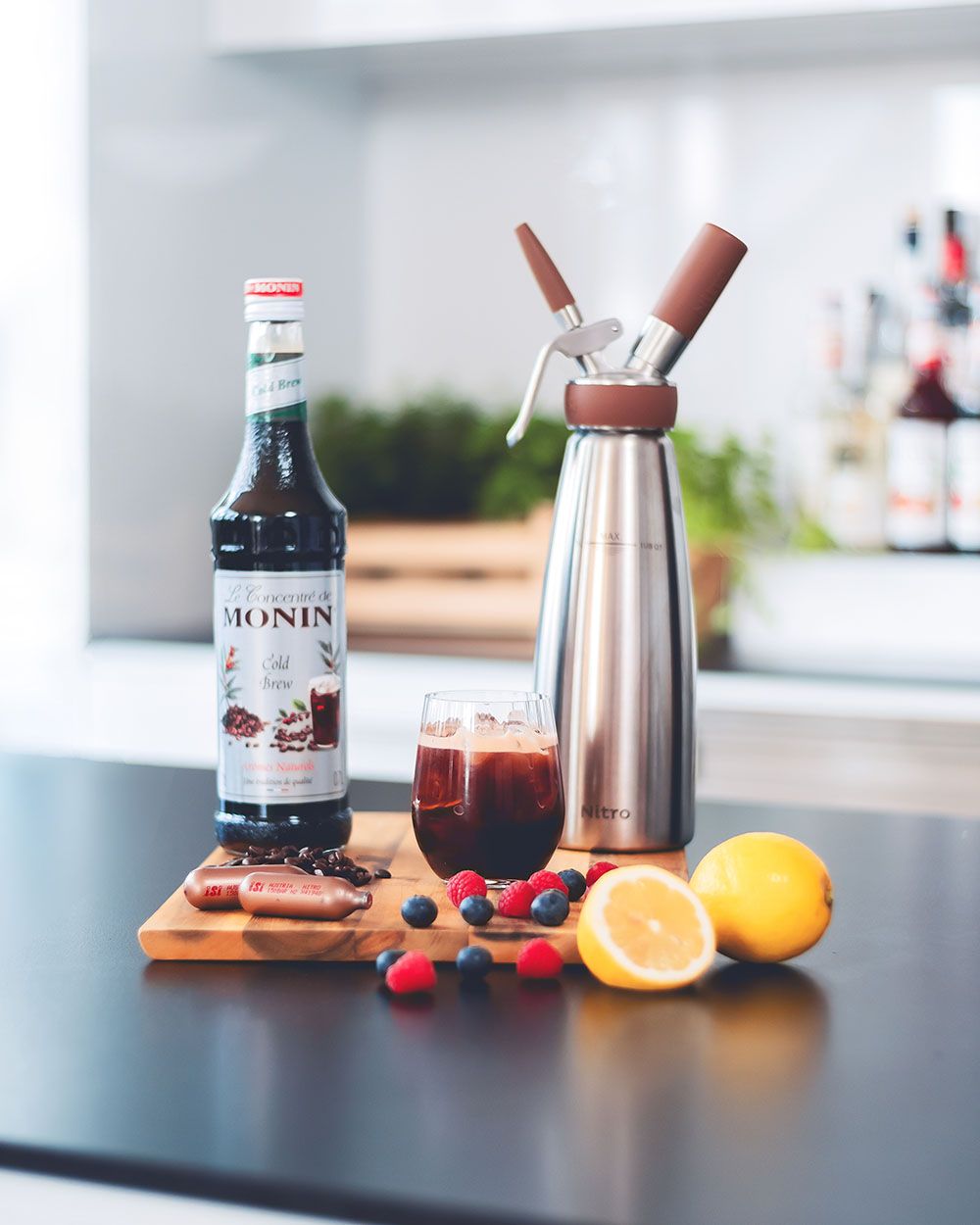 Ein Glas mit Bramble, daneben eine Flasche Monin Sirup, Beeren, iSi Charge Kapseln, Zitronen und ein iSi Nitro.