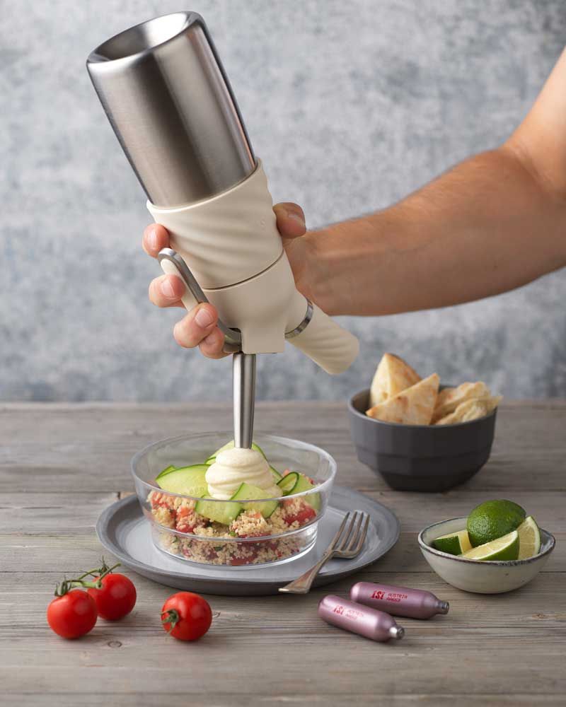 Jemand nutzt ein iSi Creative Whip, um Hummus in eine Schüssel mit Salat. Daneben Tomaten, Limetten, Brot und iSi Charge Kapseln. 