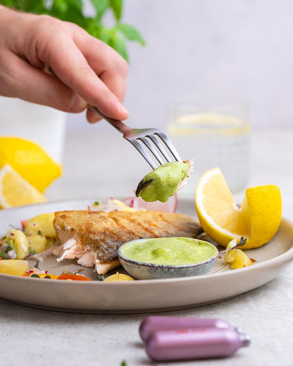Auf dem Bild wird ein Stück Fischfilet mit grüner Creme von einem Teller mit Beilagen und Zitronenscheiben mit der Gabel aufgenommen.
