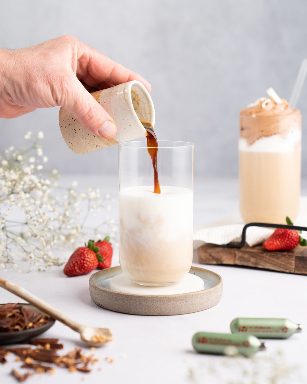 Jemand gießt Kaffee in ein Glas, daneben Erdbeeren, iSi Charge Kapseln und ein fertiges Getränk mit Nutella-Topping. 