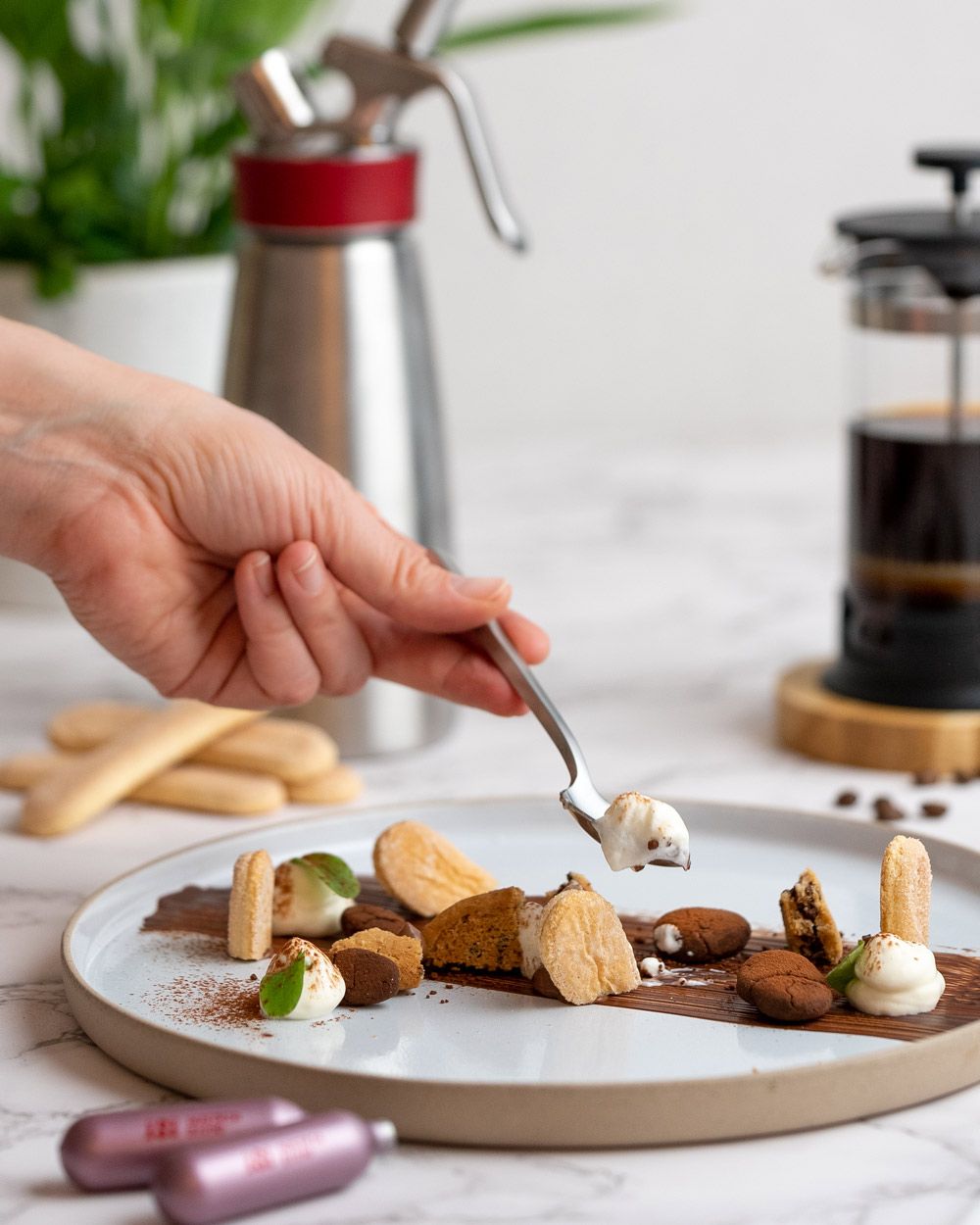 Jemand nimmt einen Löffel von einem Teller mit Tiramisu, daneben liegen iSi Charge Kapseln, im Hintergrund Biskotten, Kaffee und ein iSi Gourmet Whip.