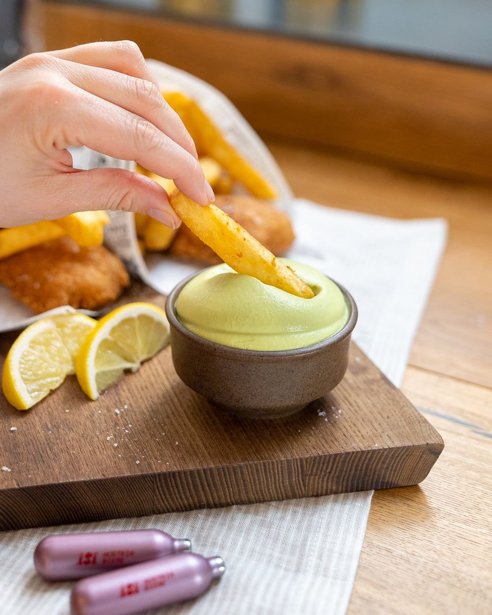 Jemand taucht ein Pommes in eine Schüssel Erbsen Espuma. Davor liegen zwei iSi Charge Kapseln, im Hintergrund zwei Tüten Fish and Chips. 