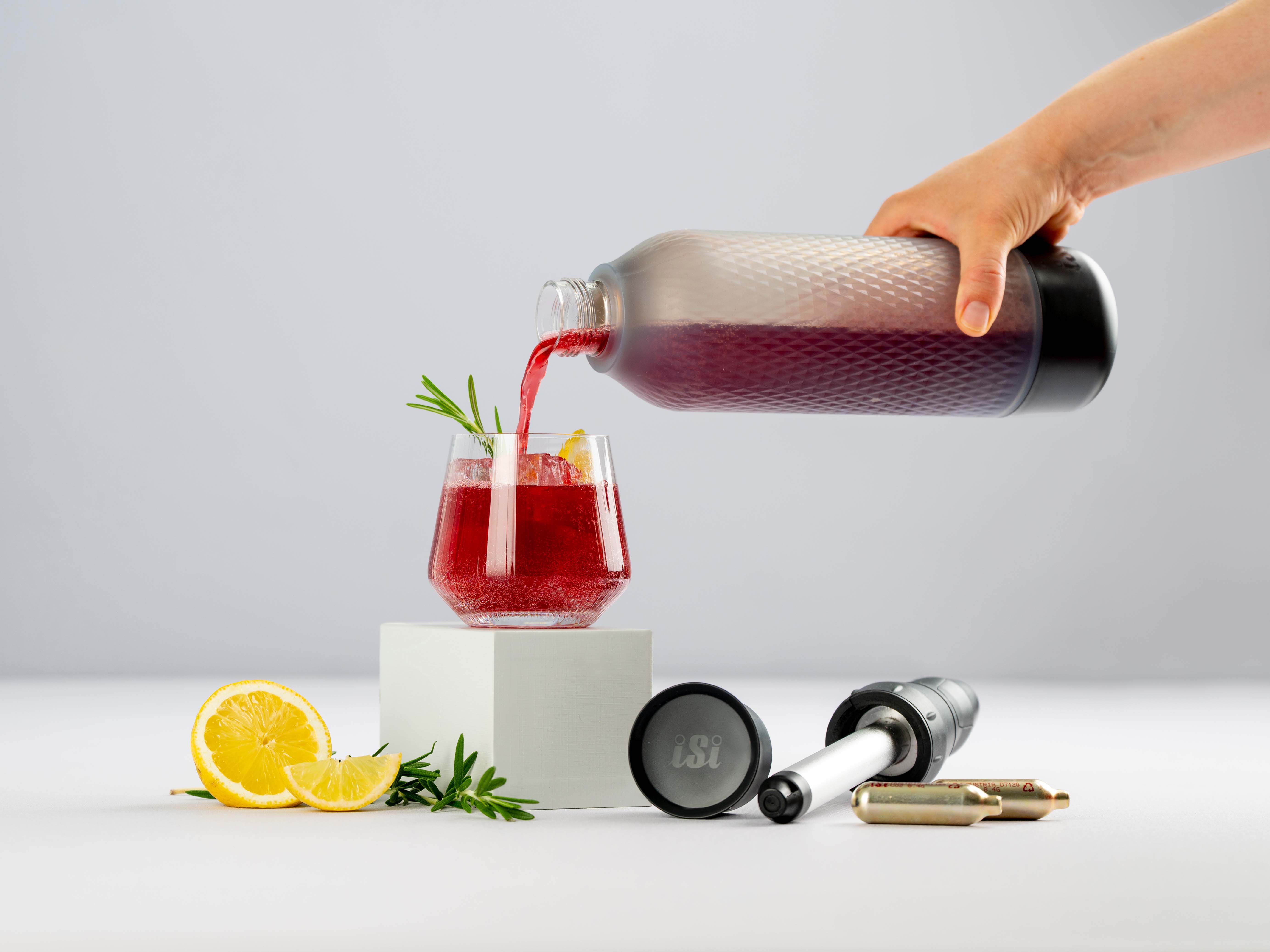 Jemand gießt aus einem iSi Twist'n Sparkle ein Johannisbeergetränk in ein Glas, daneben liegen eine Zitrone, ein Twist'n Sparkle Aufsatz und iSi Charge Kapseln.