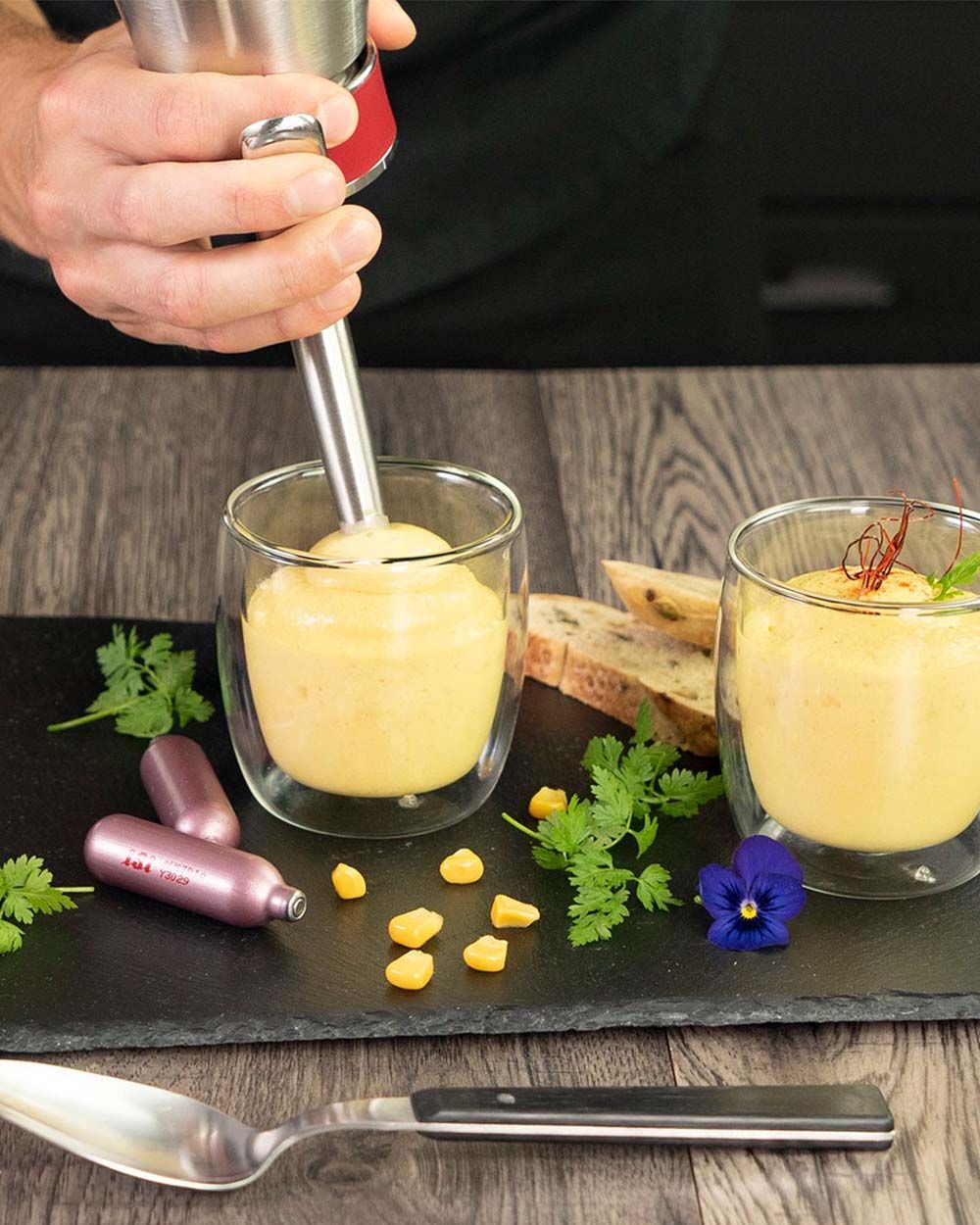 Eine Hand spritzt mit einem iSi Whip cremige Maisschaumsuppe in ein Glas, das zusammen mit Brot, Kräutern, einer Blüte und iSi Kapseln auf einer Schieferplatte angerichtet ist.