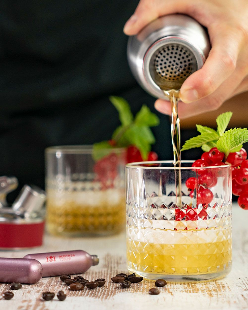 Ein Drink wird mit einem Cocktailshaker in ein Glas mit roten Johannisbeeren, Minze und Eiswürfeln eingeschenkt, während im Vordergrund iSi Kapseln und Kaffeebohnen liegen.